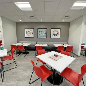 three framed artworks hanging on wall behind room of tables with red chairs